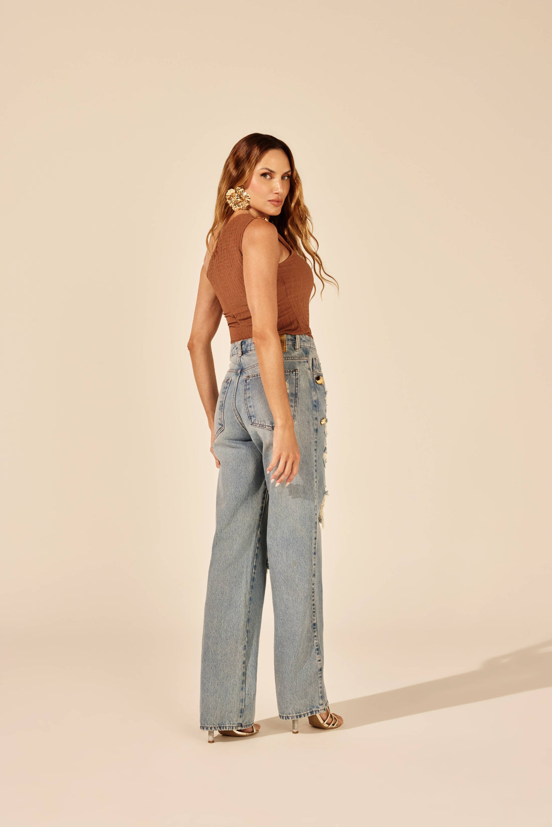 Calça reta jeans rasgada tachas douradas azul clara, look de frente com body caramelo Onça Preta