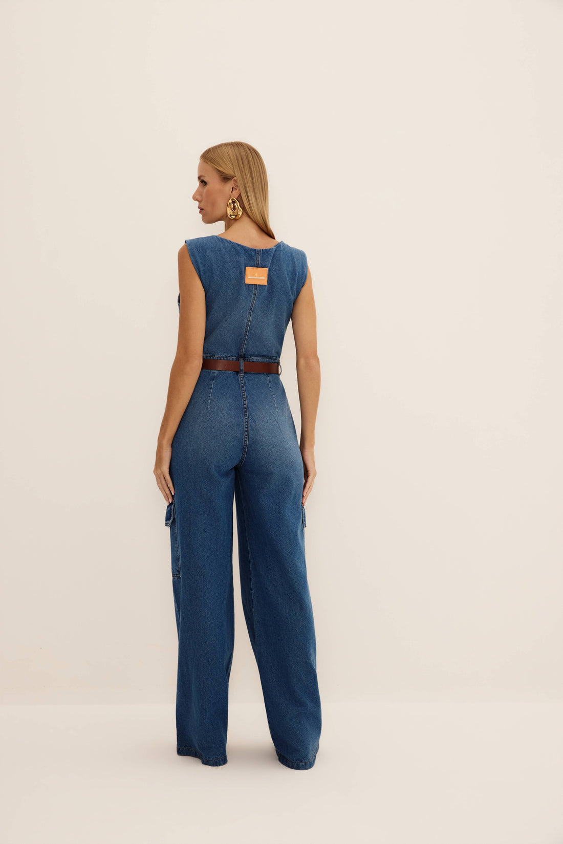 Macacão jeans reto azul regata com botões dourados, bolsos cargo e perna ampla, vista de frente com cinto