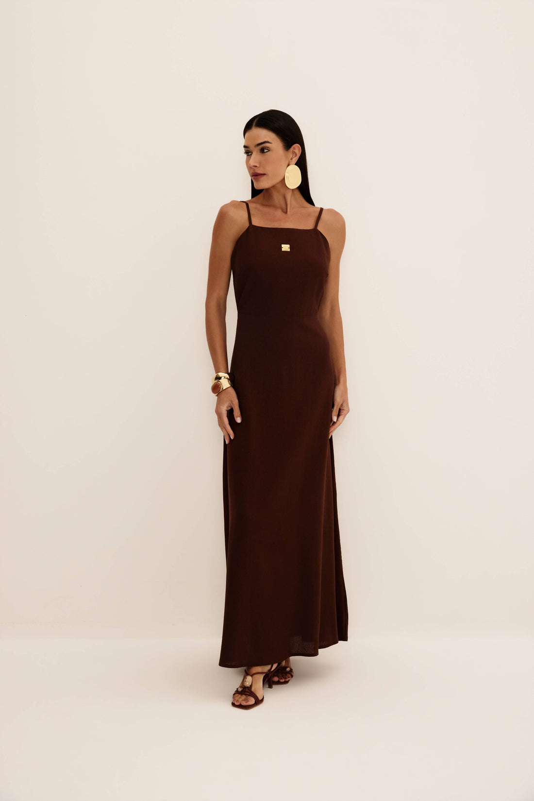 Vestido longo marrom de alças finas com decote reto, placa metálica dourada e saia fluida, vista de frente