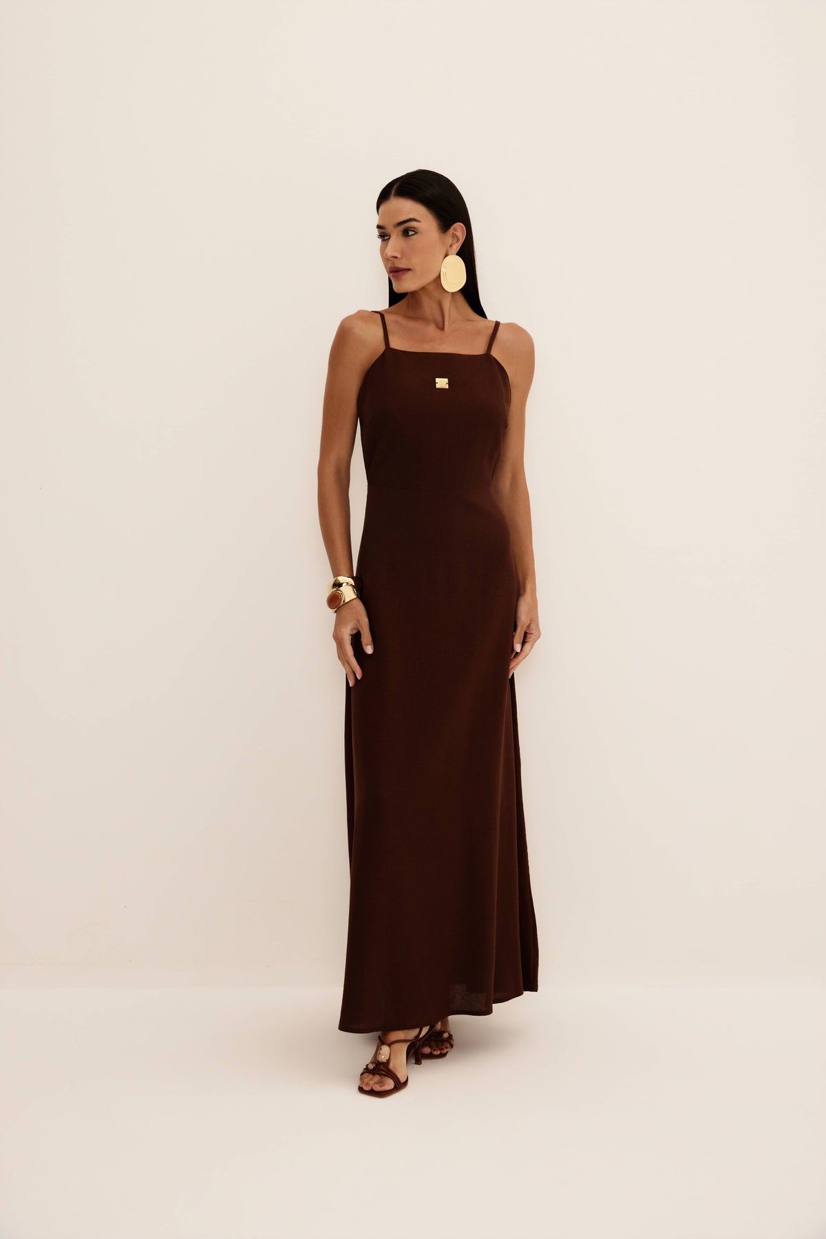 Vestido longo marrom de alças finas com decote reto, placa metálica dourada e saia fluida, vista de frente